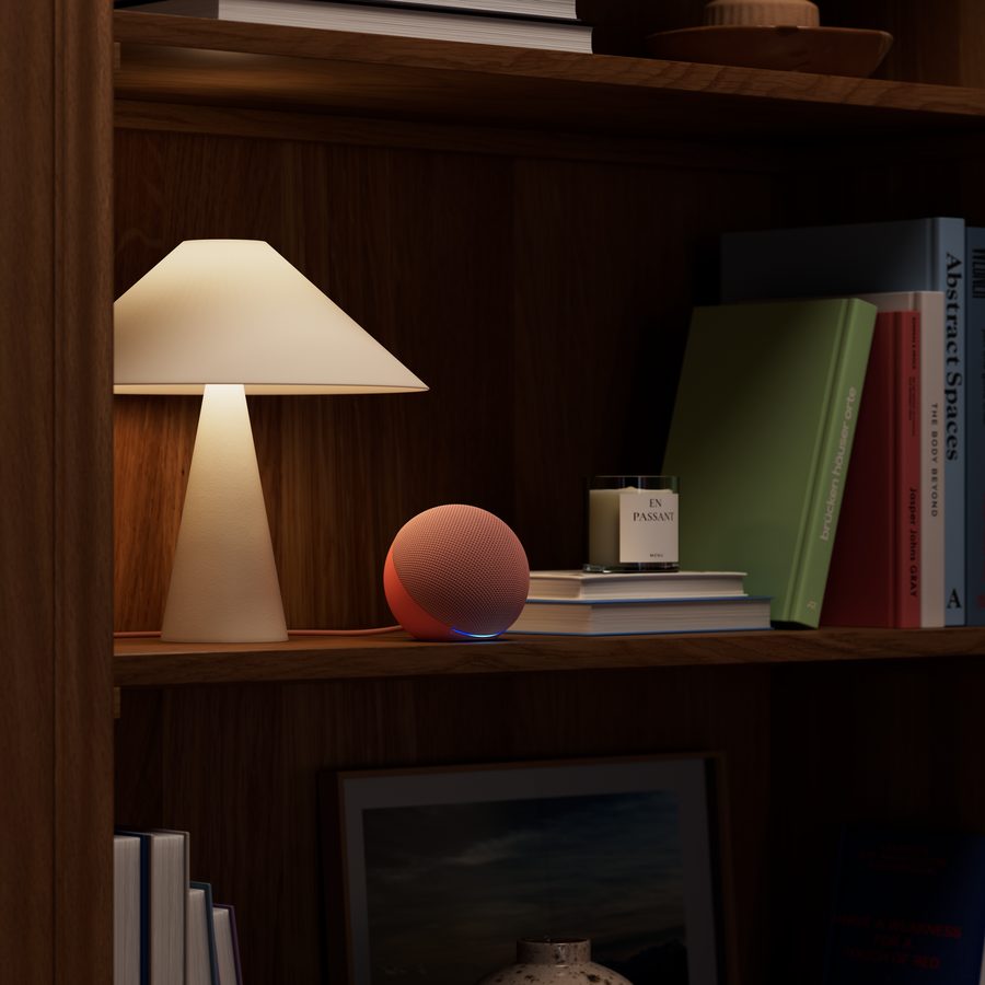 Nuevo Amazon Echo Dot esférico en color coral sobre una estantería junto a una lámpara y libros